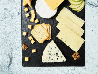 Pieces of cheese on a dark background. Cheeseboard. Sliced ​​apple and nuts on the board. Hard cheese, camembert, roquefort, emmental.