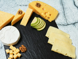 Pieces of cheese on a dark background. Cheeseboard. Sliced ​​apple and nuts on the board. Hard cheese, camembert, roquefort, emmental.