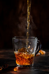 Black tea pouring into glass cup with natural steam on brown background