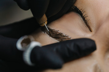 The master’s hand in a black glove holds a manipulation with a microblading needle over the model’s eyebrow, drawing hairs in macro photography.