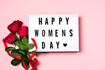 Happy women's day inscription on the lightbox, red roses with red ribbon on the pink background. 8 march concept. Top view, flat lay composition