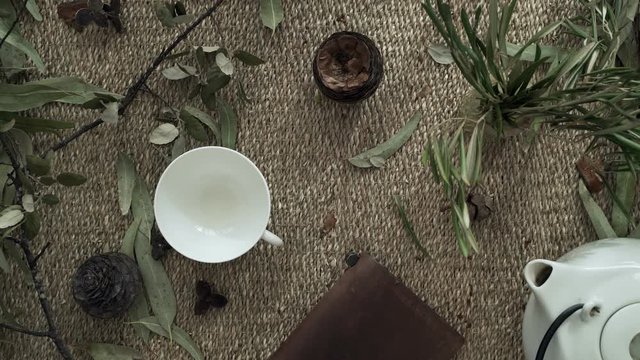 Flatlay with wicker mat, dry plants, a notebook, teapot and a cup of herbal tea. Close-up of female hands pouring tea. Tea ceremony. Drinking tea. Natural colors. Breakfast. Eco-friendly aesthetics.