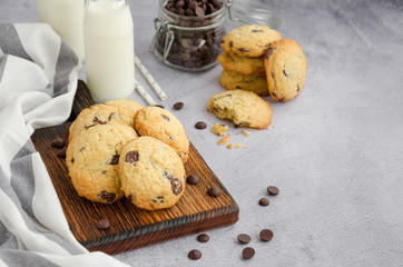Traditional American homemade cookies with chocolate chips on an old wooden board on a grey background with bottles of milk and straw. Horizontal orientation. Copy space