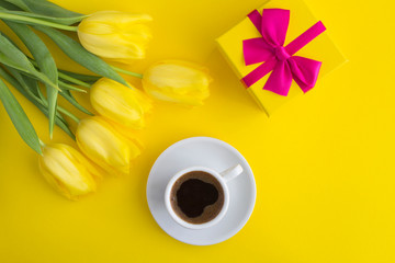 Black coffee, gift with pink bow and yellow tulips  on the yellow background. Top view. Copy space.