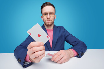 Portrait of serious young gambler in jacket sitting at table against blue background and showing ace card