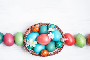 Bright multi-colored Easter eggs in a basket and spring flowers on a white wooden surface, top view. Easter background. 	