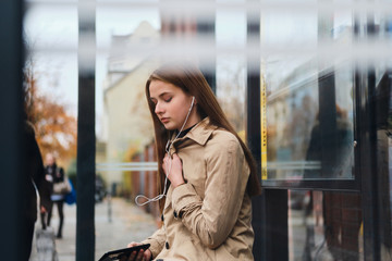 Fototapeta premium Beautiful girl in listening music in earphones dreamily waiting public transport on bus stop