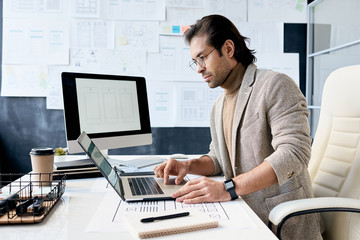 Fototapeta na wymiar Horizontal shot of handsome man wearing eyeglasses sitting at office desk working on laptop
