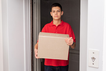 Young friendly delivery courier dressed in red T-shirt leaving the elevator holding cardboard box with order. Delivery service and logistic concept. Part-time job for teenagers and students.