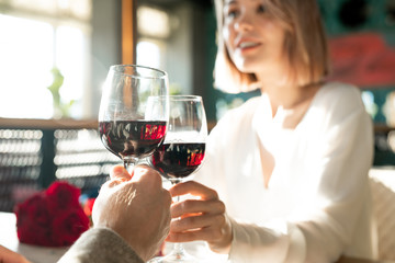 Horizontal shot of unrecognizable man and Caucasian woman sitting in front of each other clinking glasses with red wine