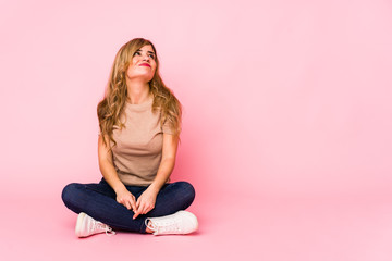Young blonde caucasian woman sitting on a pink studio dreaming of achieving goals and purposes