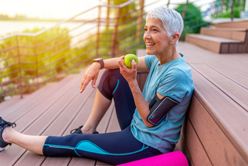 Portrait of a beautiful older woman with green healthy food after workout. Portrait of fit mature woman smiling while holding an apple and bottle of water. Sporty senior woman