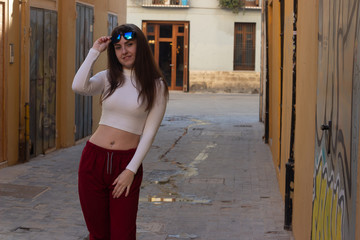 attractive caucasian young woman model looks at camera and raising stylish sunglasses, in the city, white top and red pants. Place for your text in copy space.
