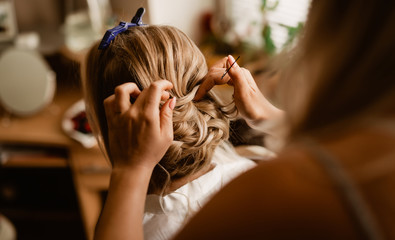 Fototapeta premium Morning hairstyle preparation of bride with hands of hairdresser. Focus on the detail of the groomed hair. Background and foreground slightly blurred.