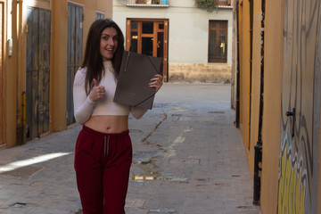 Portrait of young woman with a folder in her hands showing thumb up gesture, in the city, white top and red pants, dark air. Place for your text in copy space.