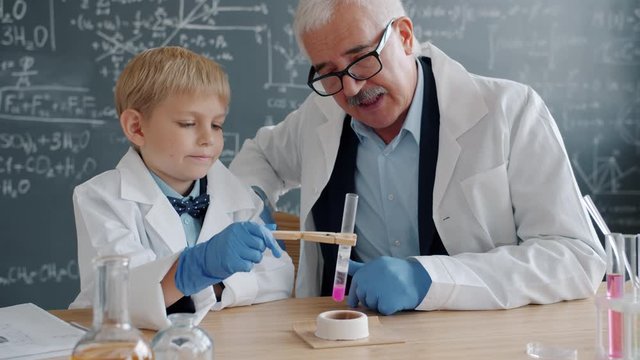 Chemists child and teacher are busy with chemical experiment in lab using test tube and burner watching reaction, blackboard with formulas is in background.