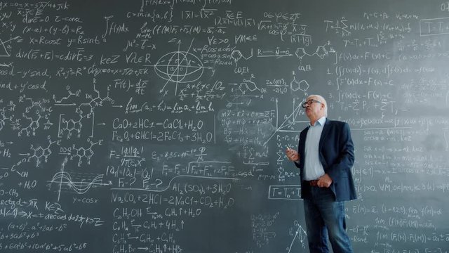 Thoughtful man is walking near chalkboard wall thinking then writing formulas finding solution for scientific problem. People, science and education concept.