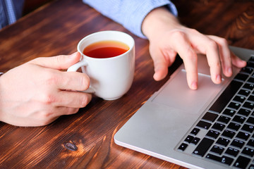 A man in a shirt is drinking tea and browsing the web in a laptop.