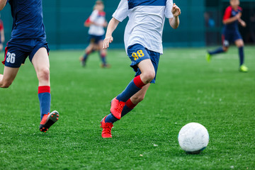 Boys in white and blue sportswear plays  football on field, dribbles ball. Young soccer players with ball on green grass. Training, football, active lifestyle for kids concept 