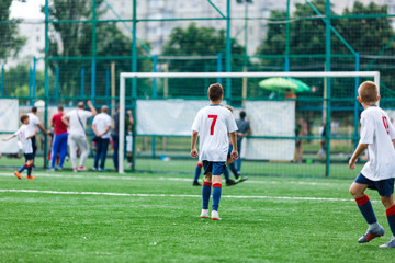 Obraz premium Boys in white and blue sportswear plays football on field, dribbles ball. Young soccer players with ball on green grass. Training, football, active lifestyle for kids concept 