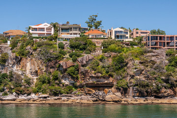 Fototapeta na wymiar Sydney, Australia - December 11, 2009: Upscale housing with red roofs on rocky cliff with green vegetation on northern shore of bay under blue sky and behind green bay water.