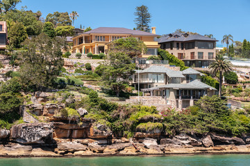 Sydney, Australia - December 11, 2009: Upscale housing with gray roofs on rocky slope with green vegetation on northern shore  of bay under blue sky and behind green bay water.