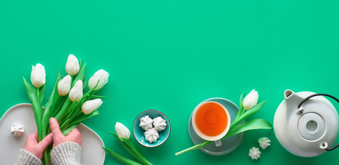Geometric panoramic flat lay with hands. Cup of black tea, tea pot, sweets and white tulips on green with copy-space. Valentine day, Mother's day, international women day March 8. Trendy flat lay.