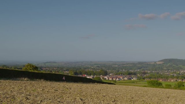 A daylight extreme long shot of a man carrying a backpack hiking down a dried plain with sparse grass towards a township.
