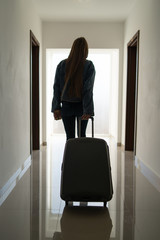 Silhouette of young good looking girl back view female woman walking in the bright lobby hall against the window at the hotel or motel dragging her suitcase luggage posing full length