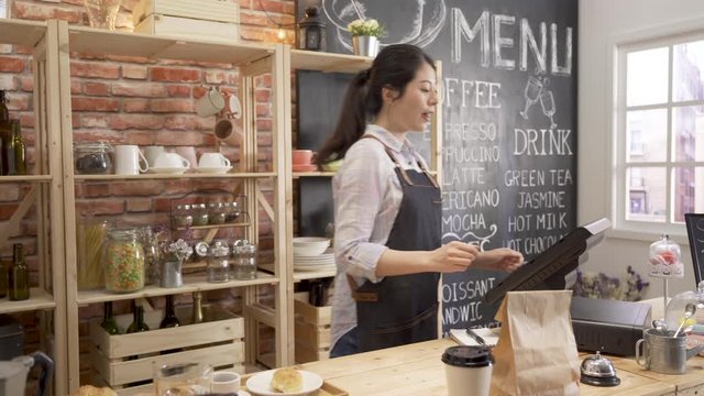 Attractive Barista In Apron Putting Paper Cup And Bag On Bar Counter Table And Man Customer Takeaway By Himself In Coffee Shop. Young Girl Waitress Waving Hands Saying Goodbye And Working On Cashbox