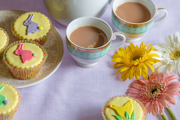 Tea time with pretty colorful yellow Easter cupcakes with pastel icing shapes of Easter bunny rabbits and chicks and Easter flowers
