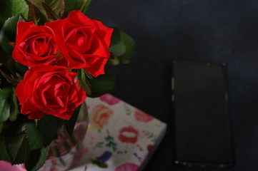 Bouquet red roses flower in glass vase on dark background and black smartphone no focus, gift box