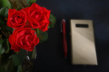Bouquet red roses flower in glass vase on dark background and pen at the gold smartphone no focus, gift box