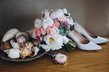 On a wooden table are white shoes, next to them is a wedding bouquet of flowers and greens, a glass box for engagement rings and a vase with shells