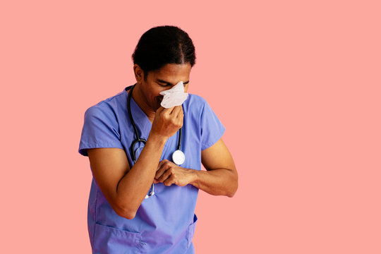 Studio Portrait Of A Sick Male Doctor Or Nurse Wearing Blue Scrubs And Stethoscope Blowing Nose