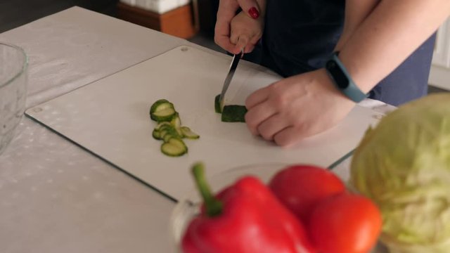 Close-up of a mother with a little daughter in identical aprons preparing a vegetable salad together, they hold together by a knife and cut a fresh cucumber. Slow motion.
