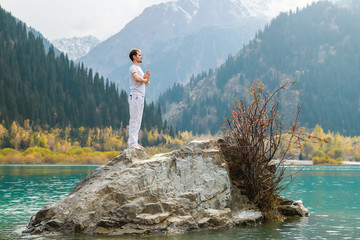 A man in a Samasthiti pose on a stone among a mountain lake.