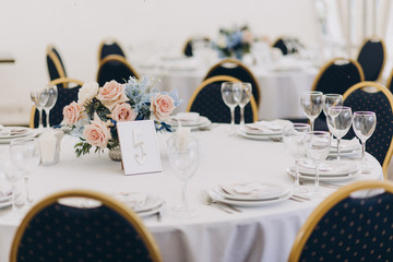 holiday tables are decorated with compositions of flowers and greenery, on the tables are tablecloths, plates with napkins and postcards, glasses, candles and cutlery