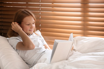Cute preteen girl reading book in bed near window