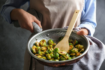 Woman with frying pan of roasted Brussels sprouts on grey background, closeup