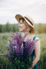 A beautiful romantic woman smiles joyfully and looks into the distance in a blue dress and hat in a flower field with a huge bouquet of purple lupine flowers. Soft selective focus.