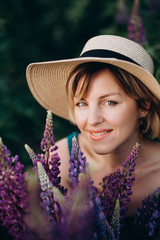 A beautiful romantic woman smiles joyfully in a blue dress and hat sits in a flower field with a huge bouquet of purple lupine flowers. Close-up. Soft selective focus.