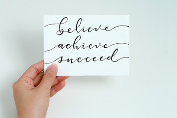 Female hand holding white card with calligraphy lettering Believe Achieve Succeed