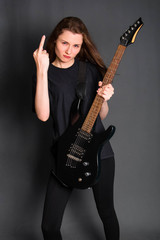 Angry rock girl in black clothes holds an electric guitar in her hands and shows the middle finger, gesture fuck off. Studio photo on a gray background.