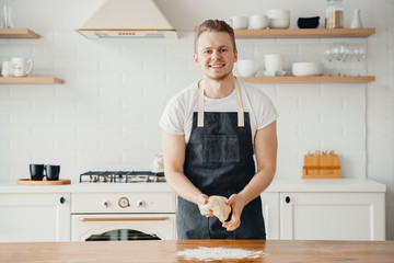 Male chef cook baker smiles, holds fresh dough in hands in apron, light background kitchen