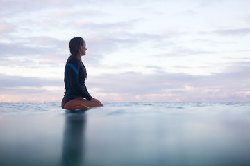 Woman sitting on surfboard looking out to sea horizon at sunset