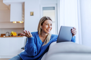 Beautiful caucasian blond woman dressed in blue sweater sitting on sofa in living room and having video call over tablet. - obrazy, fototapety, plakaty