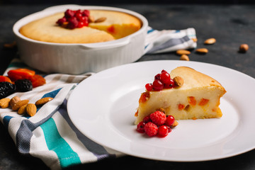 Freshly baked homemade cottage cheese pie with dried apricots, almonds and raspberries. A piece on a white plate decorated with berries and Cutlery side View on a dark background