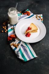 Freshly baked homemade cake with cottage cheese and dried apricots, almonds and raspberries and a glass of milk. A piece on a white plate decorated with berries and Cutlery side View on a dark backgro