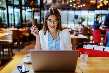 Good-looking positive fashionable charming brunette sitting in cafe and using benefits of new credit card. She is using laptop for online shopping.